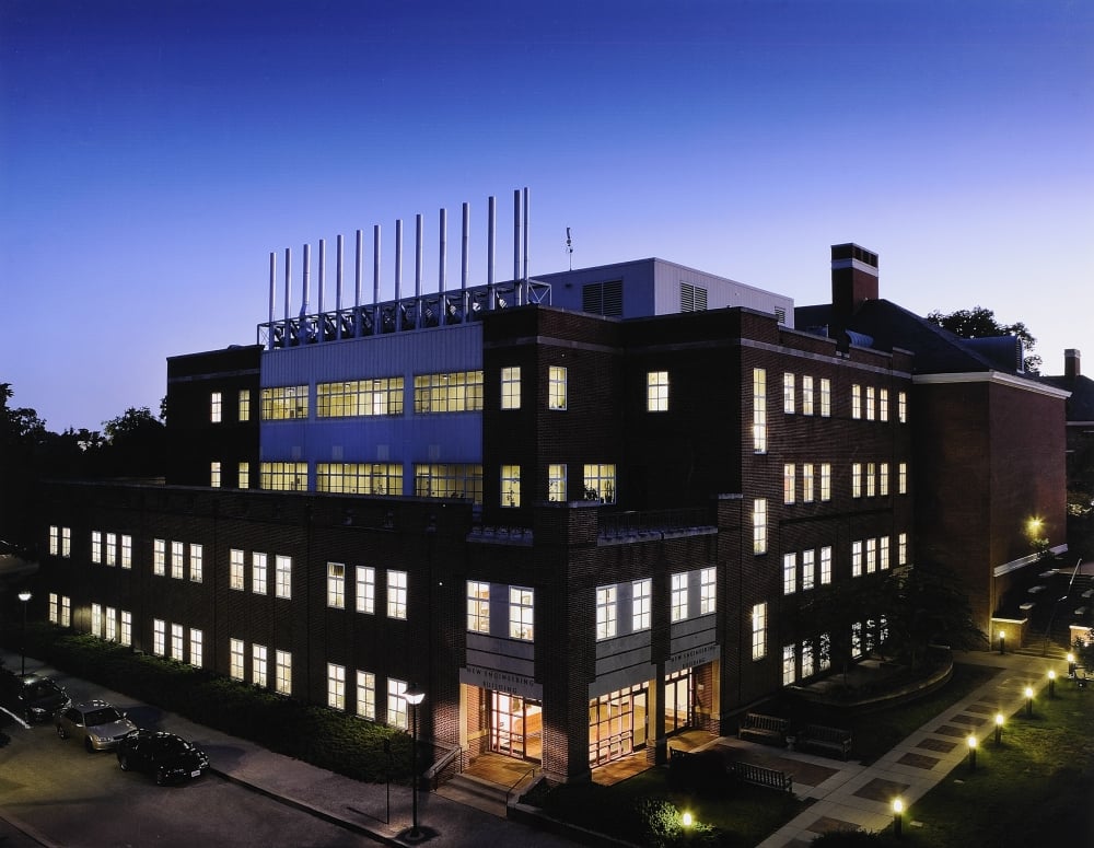 Four-story rectangular red brick building at dusk with all the lights on. There is a row of outdoor lights on the right-side of the building along the sidewalk and three cars parked along the left-side of the building.